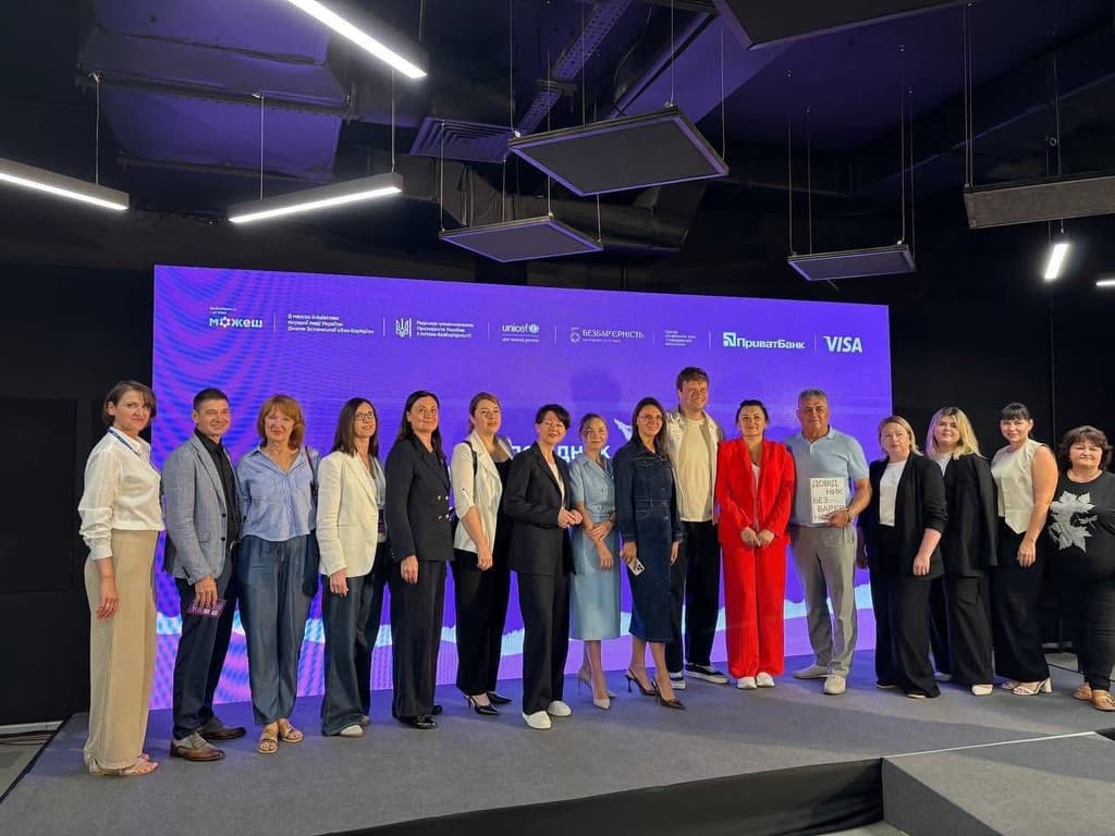 Fifteen individuals stand on a stage in front of a large digital screen displaying Cyrillic text and logos of HORIZONT, UNICEF, Belarusbank, and VISA. One person holds a certificate or plaque, suggesting an award or recognition. The group is dressed in a mix of business and casual attire, indicating a semi-formal event. The indoor venue features modern lighting and ceiling fixtures, creating a professional and celebratory atmosphere.