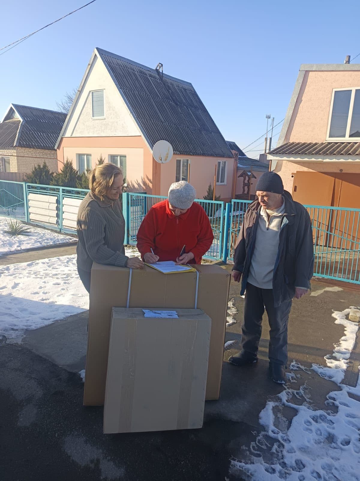 Троє людей стоять на вулиці на бруківці з плямами снігу. Вони зібралися навколо двох великих картонних коробок, поставлених одна на одну. Людина в червоній куртці та сірій в’язаній шапці пише на планшеті чи аркуші, покладеному на коробки. Інший учасник у зеленій куртці допомагає або спостерігає, третій у темному одязі та шапці стоїть поруч. На задньому плані видно житлові будинки пастельних кольорів та синю металеву огорожу. Сцена передає процес доставки чи отримання товарів із оформленням документів.