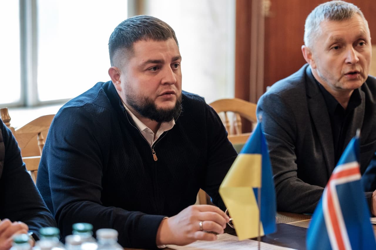 Двоє чоловіків у темних костюмах сидять за столом під час офіційної зустрічі, перед ними — маленькі прапори України та Ісландії. Один із чоловіків — у білому сорочці під темним светром, з бородою. На столі — пляшки з водою та документи, а дерев’яні стільці й панелі створюють атмосферу урядового або офіційного приміщення. Сцена передає дипломатичну або партнерську взаємодію між представниками України та Ісландії.