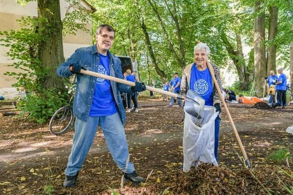 The image shows two individuals participating in a community clean-up activity in a wooded area. Both individuals are wearing blue shirts with a white circular logo and are using tools to collect leaves and debris into a large white bag. One person is holding a rake, while the other is holding a shovel. In the background, there are more people involved in the clean-up, some of whom are also wearing blue shirts. There are trees and a building visible in the background, and the ground is covered with leaves. This image is interesting and relevant as it depicts community involvement and environmental stewardship.