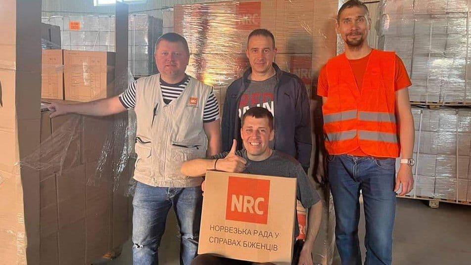 Four men posing in a warehouse among boxes of humanitarian aid. One of them, sitting in a wheelchair, holds a box with the NRC (Norwegian Refugee Council) logo, and everyone is smiling.