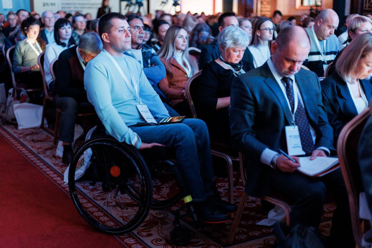 The image shows a group of people seated in an auditorium or conference room. The individuals are attending an event, possibly a conference or seminar, as indicated by the name tags and lanyards they are wearing. One person in the foreground is using a wheelchair and has a tablet on their lap. Another person next to them is taking notes in a notebook. The setting appears formal, with attendees dressed in business or casual attire. The room is well-lit, and the audience is focused on the event.