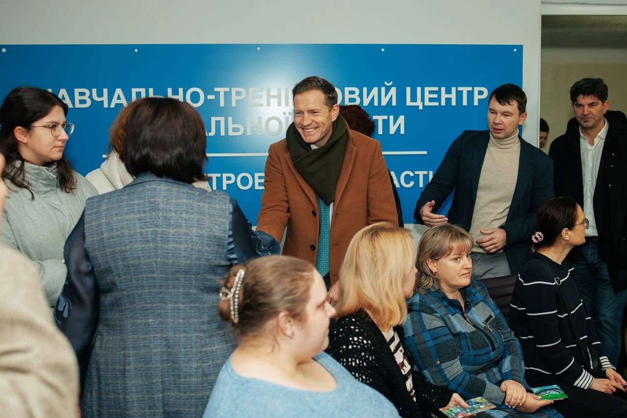 A group of people gathers indoors near a blue banner reading “Training Center for Social Work, Dnipropetrovsk Region.” A man in a brown coat and green scarf smiles and shakes hands with a woman. Others are seated or standing, engaged in conversation or listening. The scene reflects a welcoming or introductory moment at a social work-related event.