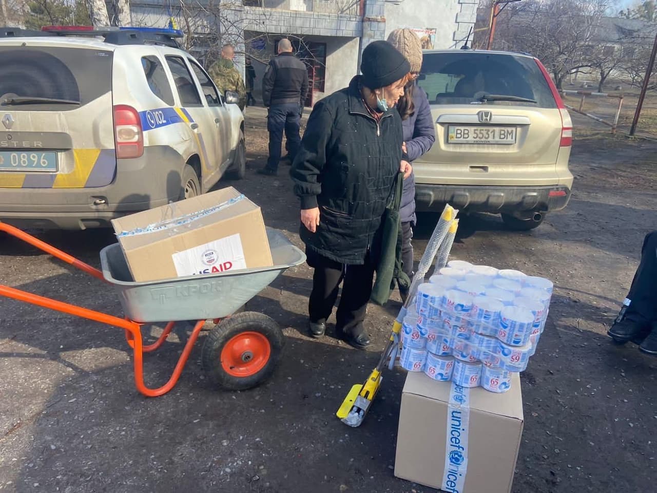Сцена розподілу гуманітарної допомоги на вулиці. На передньому плані тачка з картонною коробкою з логотипом «USAID». Поруч велика коробка з написом «UNICEF», на якій складено кілька паків консервів із маркуванням «65». До коробки притулений жовто-сріблястий інструмент чи пристрій. Біля допомоги стоять кілька людей у зимовому одязі, серед них літня людина в чорному пальті та капелюсі. На задньому плані видно поліцейський автомобіль із номером «0896» та бежевий Honda SUV із номерним знаком «BB 5531 BM». Сцена демонструє міжнародну підтримку та розподіл допомоги в житловому районі.