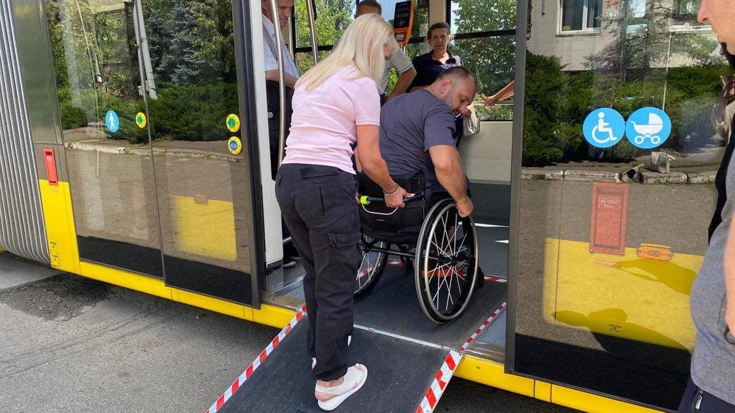 A person in a wheelchair is assisted by another individual while boarding public transportation via an accessibility ramp. The ramp extends from the vehicle door to the ground, enabling easy access. Accessibility symbols for wheelchairs and strollers are displayed on the door. Several people inside the vehicle are visible, observing or assisting. The scene highlights the importance of accessible public transport and support for individuals with mobility challenges.