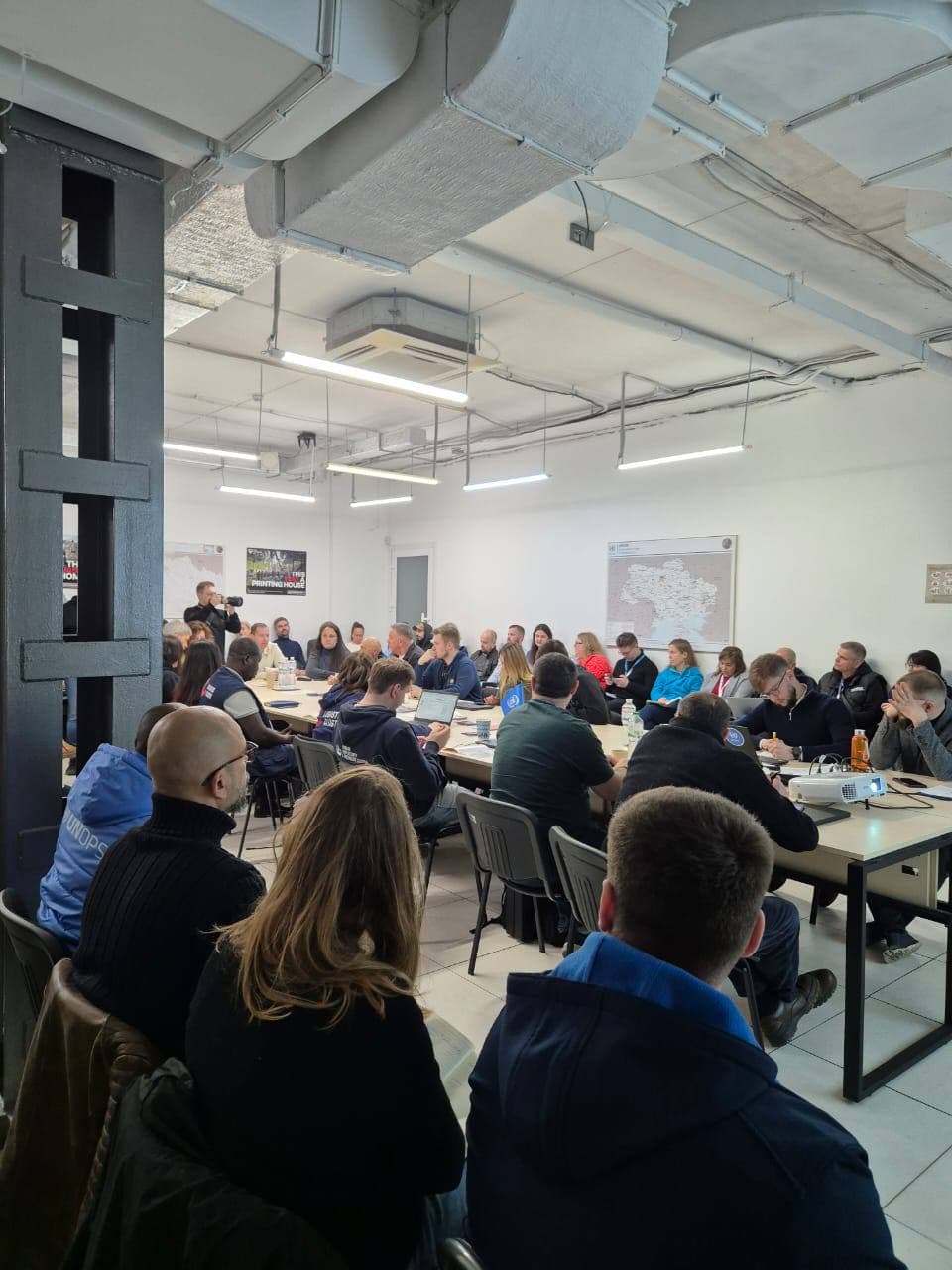 Яскраво освітлений конференц-зал із великою групою людей, які сидять за кількома столами. Приміщення має індустріальний стиль із відкритими трубами та вентиляційними каналами. Учасники ведуть дискусії або переглядають документи, створюючи професійну та кооперативну атмосферу. На стінах розміщені постери, серед яких один схожий на карту.