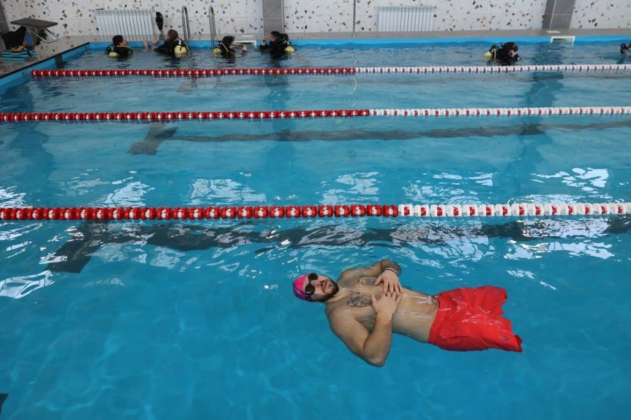 Критий басейн із кількома доріжками, розділеними червоно-білими буйками. На передньому плані людина у червоних плавках, рожевій шапочці та окулярах лежить на спині у воді. На задньому плані кілька осіб у спорядженні для дайвінгу стоять біля краю басейну, готуючись до занурення чи тренування. Стіни облицьовані плиткою, видно радіатори. Сцена поєднує рекреаційне плавання та навчання дайвінгу в одному просторі.