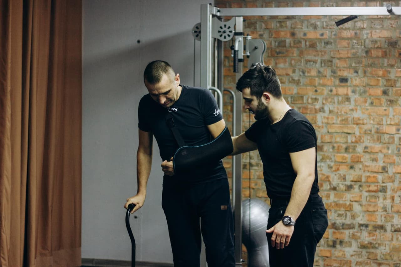 Two men, one of whom is trying to walk with the support of a crutch, while the other is helping him.