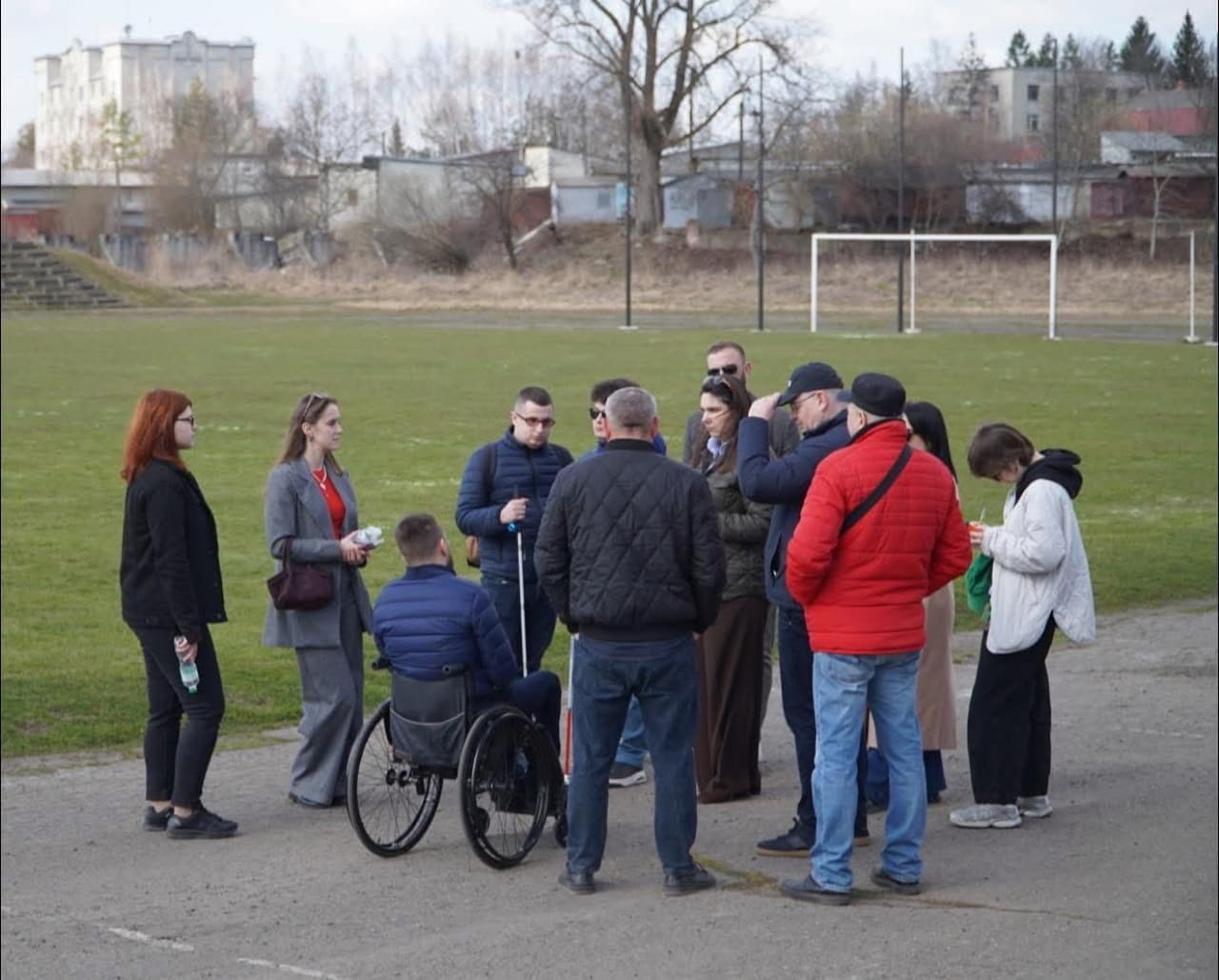 На зображенні група людей зібралася на відкритому майданчику біля футбольного поля. Один із учасників сидить у інвалідному візку, інші стоять поруч, спілкуються або слухають. На задньому фоні видно ворота для футболу, трав’яне поле, будівлі та дерева без листя, що вказує на холодну пору року. Люди одягнені у повсякденний та напівофіційний одяг, деякі тримають пляшки з водою чи блокноти. Атмосфера нагадує зустріч громади або інклюзивний захід.