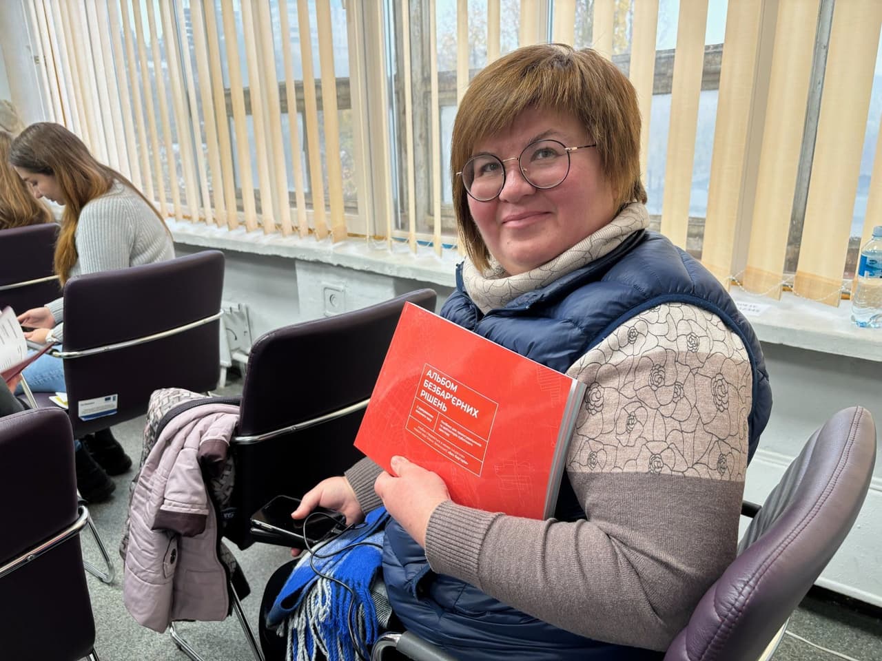 Учасник семінару тримає червону книгу з назвою «Алгебра безопасных решений» російською мовою. Кімната оформлена як навчальний простір: люди сидять на стільцях, читають матеріали, на підвіконні — пляшка води. Атмосфера сприяє навчанню та обговоренню рішень в умовах невизначеності.