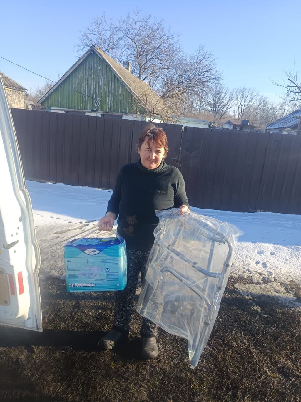 Людина стоїть на вулиці біля білого автомобіля з відчиненими дверима. У лівій руці тримає упаковку підгузків для дорослих TENA Slip, у правій — складний ходунок, загорнутий у прозору плівку. На задньому плані видно коричневий паркан, зелену будівлю з похилим дахом та дерева без листя. Земля вкрита снігом, що свідчить про зимову пору. Сцена демонструє доставку чи транспортування необхідних медичних засобів для догляду за людьми похилого віку або людьми з інвалідністю.