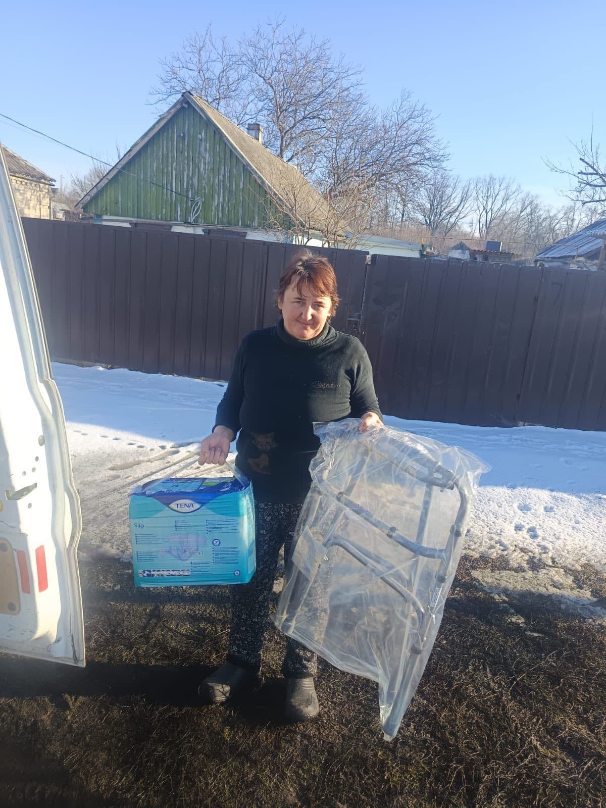 Людина стоїть на вулиці біля білого автомобіля з відчиненими дверима. У лівій руці тримає упаковку підгузків для дорослих TENA Slip, у правій — складний ходунок, загорнутий у прозору плівку. На задньому плані видно коричневий паркан, зелену будівлю з похилим дахом та дерева без листя. Земля вкрита снігом, що свідчить про зимову пору. Сцена демонструє доставку чи транспортування необхідних медичних засобів для догляду за людьми похилого віку або людьми з інвалідністю.