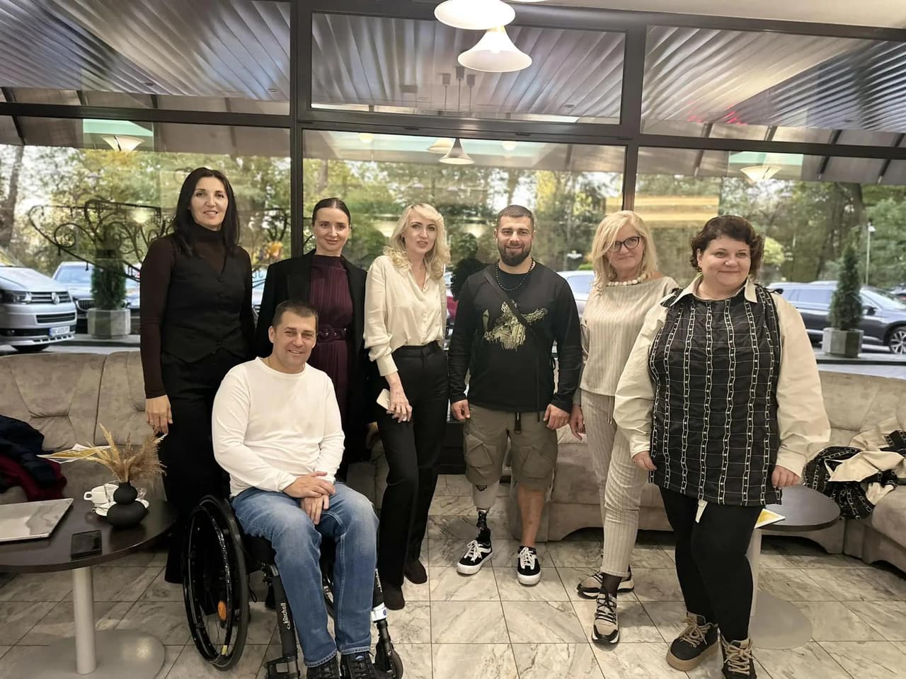 Група учасників публічної дискусії, зокрема представники ГС ЛАООІ, включаючи голову організації Миколу Надулічного, позують для фото на фоні інтер’єру з панорамними вікнами. Один із учасників у інвалідному візку, інший — із протезом ноги. Усі посміхаються, створюючи атмосферу дружності та співпраці.