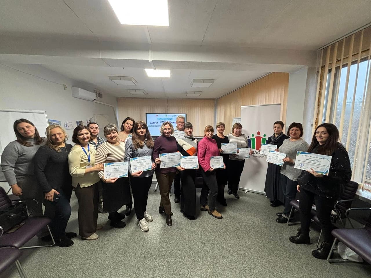 Group photo of fifteen people in a room with white walls, chairs, and a flip chart. Most hold certificates. In the background, a screen displays the text “Protection. Creation of a Safe Space” along with logos, including UNHCR. A colorful banner with stylized human figures is visible on the right. The scene captures the completion of a training or educational program focused on safety, inclusion, or community support.