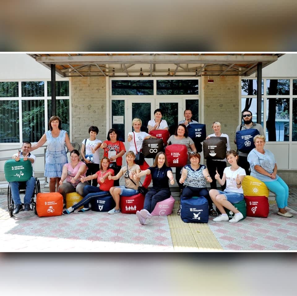 A group photo of event participants outdoors. People hold cushions featuring Sustainable Development Goals symbols. Everyone is smiling and showing a positive attitude.