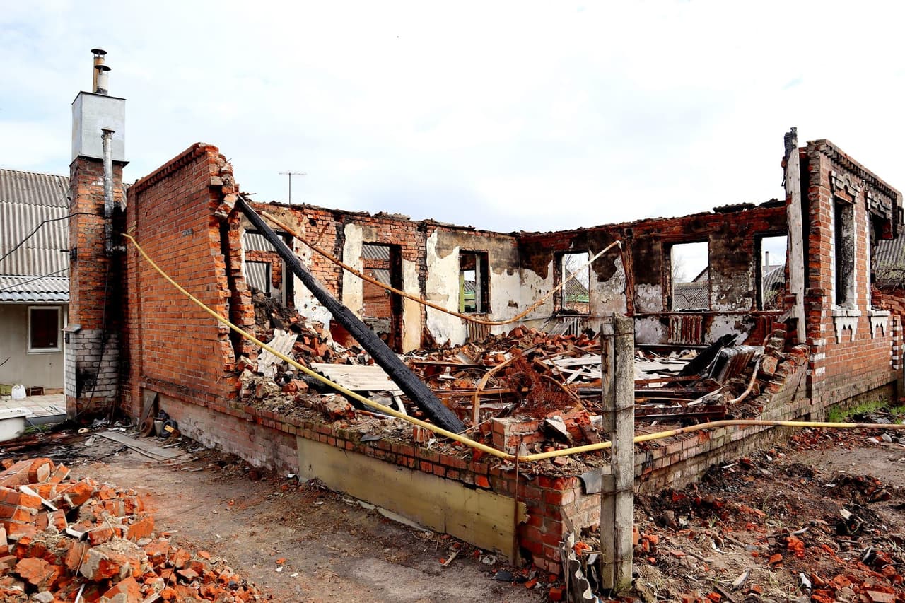 Зруйнований будинок з обгорілими стінами та заваленим дахом, залишки будівельних матеріалів на землі.