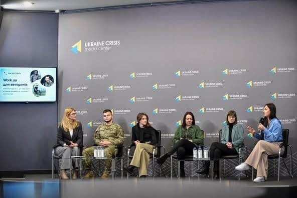 На зображенні зображено панельну дискусію в Українському кризовому медіацентрі. На сцені сидять шість осіб: п'ять жінок і один чоловік у військовій формі. На фоні видно логотип Українського кризового медіацентру, який повторюється. Ліворуч розташований екран із текстом "Work.ua для ветеранів" і зображеннями людей. Перед учасниками на столі стоять пляшки з водою. Світлина відображає захід, присвячений обговоренню питань, пов'язаних із ветеранами.