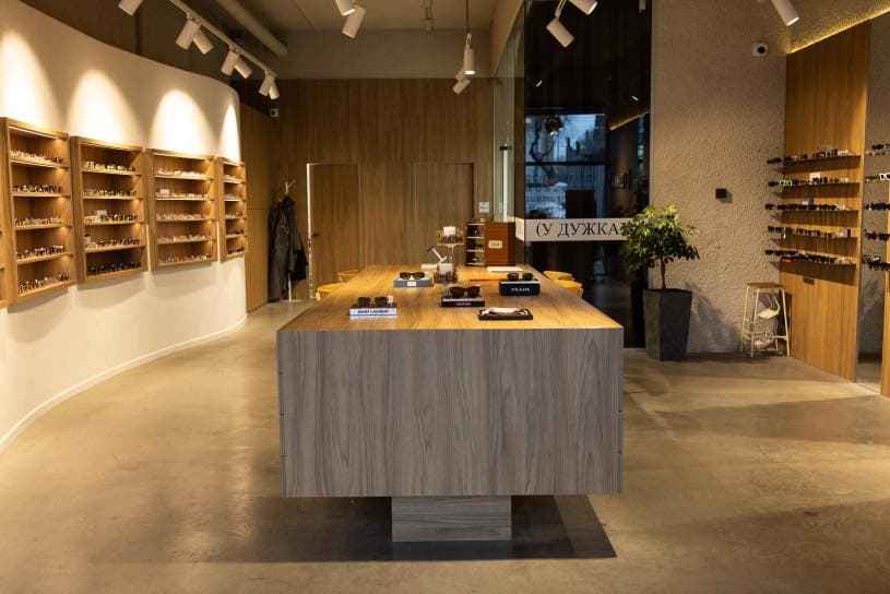 Interior of a modern eyewear store. A central wooden table displays neatly arranged eyeglasses and sunglasses, including brands like Prada and Gentle Monster. The left wall features multiple shelves with a wide selection of frames, while the right wall has additional shelves and a potted plant. Warm spotlighting highlights the products. In the background, a coat hangs near a counter area for customer service. The space is minimalist and stylish, designed for a comfortable shopping experience.