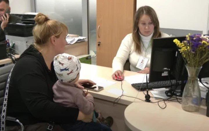 Жінка з дитиною сидить за столом навпроти працівниці офісу, яка працює за комп’ютером і має бейдж на шиї. На столі — документи та ваза з квітами. У фоні видно іншу людину, яка говорить по телефону біля принтера. Сцена відображає момент отримання адміністративних чи соціальних послуг у робочому середовищі.