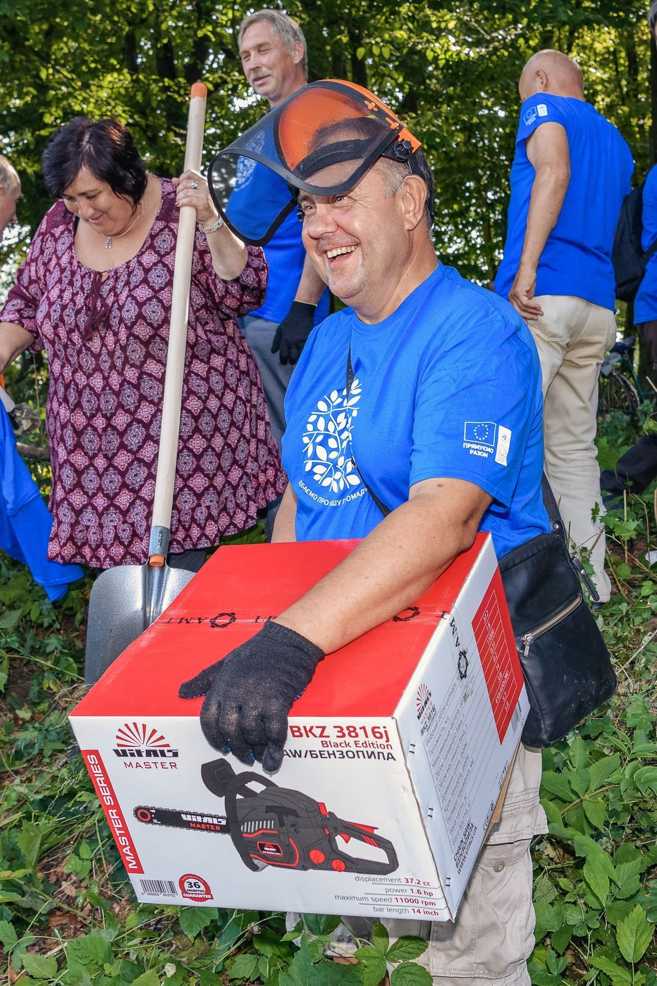 The image shows a group of people engaged in an outdoor activity, possibly a community service or volunteer event. One person in the foreground is holding a large box containing a chainsaw, specifically a "BKZ 3816j Black Edition" from the "Master Series" by the brand "VIKING MASTER." The box details include specifications such as displacement (37.2 cc), power (1.6 hp), maximum speed (11000 rpm), and bar length (14 inches). The people in the image are wearing blue shirts with a logo and text, and one person is holding a shovel. The setting appears to be a wooded area.