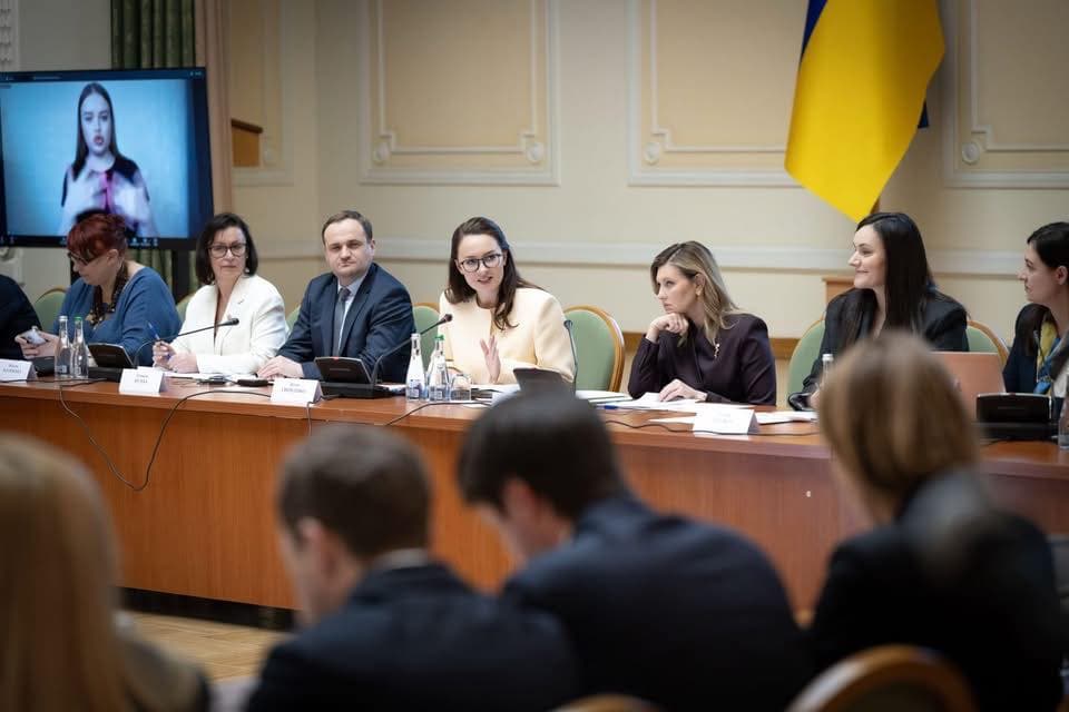 Формальна зустріч у добре освітленій залі з декоративними стінами та українським прапором на задньому плані. За довгим дерев’яним столом сидять кілька людей у діловому одязі, перед ними таблички з іменами, мікрофони та документи. На екрані позаду видно жінку, яка використовує жестову мову, що свідчить про забезпечення доступності для людей із порушенням слуху. Атмосфера офіційна та серйозна, учасники уважно слухають і обговорюють.