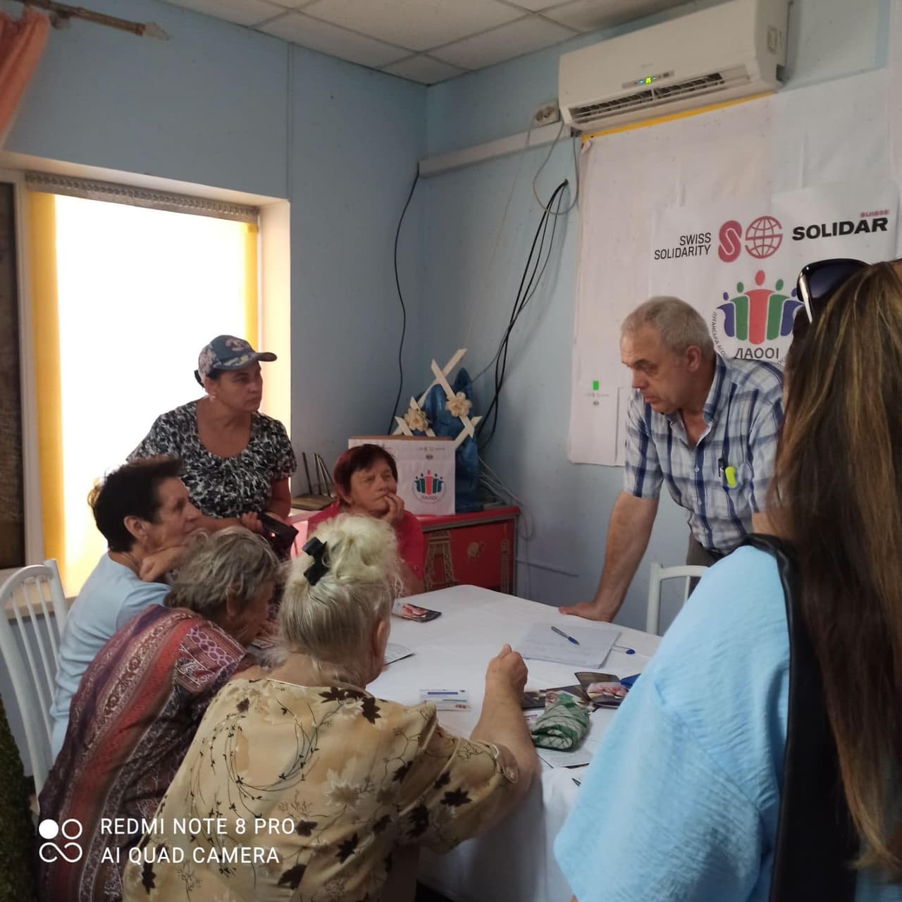 Чотири жінки сидять за столом, одна жінка стоїть, а навпроти біля столу стоїть чоловік, ще одна жінка стоїть спиною.