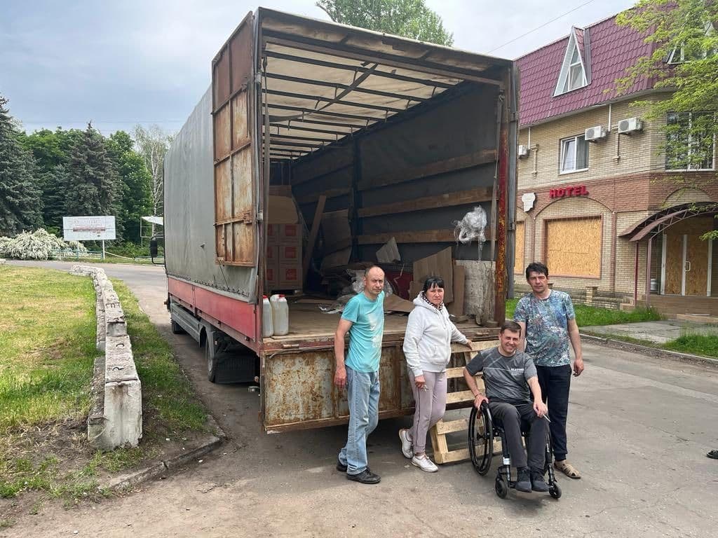 Four people stand next to an open cargo truck parked on a street. One person is seated in a wheelchair. Inside the truck are cardboard boxes and plastic containers. In the background is a building with a red roof and a “HOTEL” sign, with boarded-up windows and doors. Trees, grass, and a concrete barrier line the road. The scene suggests a loading or unloading moment, possibly related to humanitarian aid or community support.