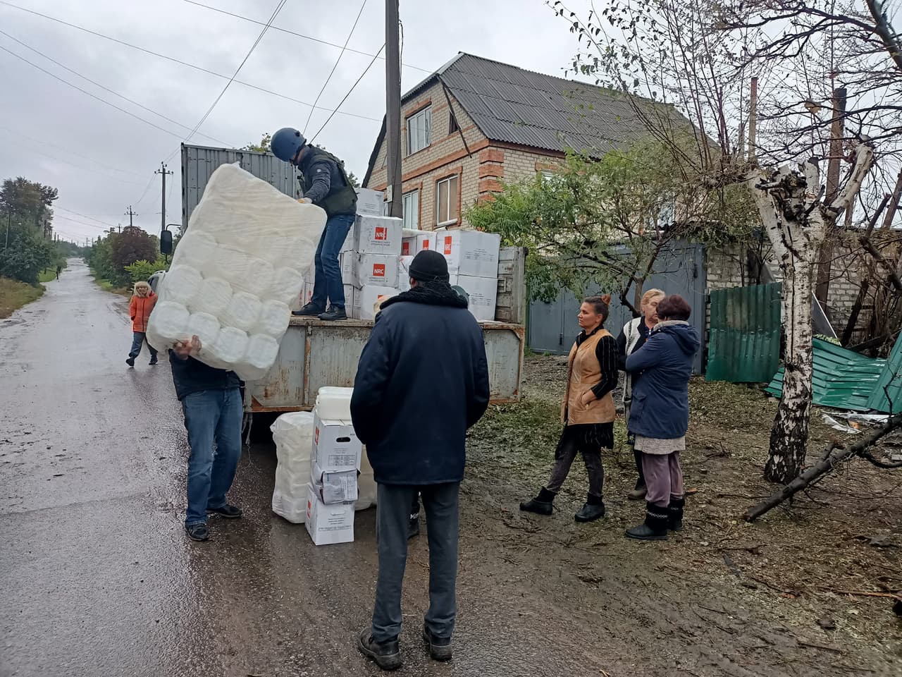 Humanitarian aid in a conflict zone. Volunteers are unloading a truck, handing over large packages and boxes to people gathered nearby. In the background, a damaged fence and houses are visible. The gloomy, rainy weather highlights the difficult conditions faced by the residents.