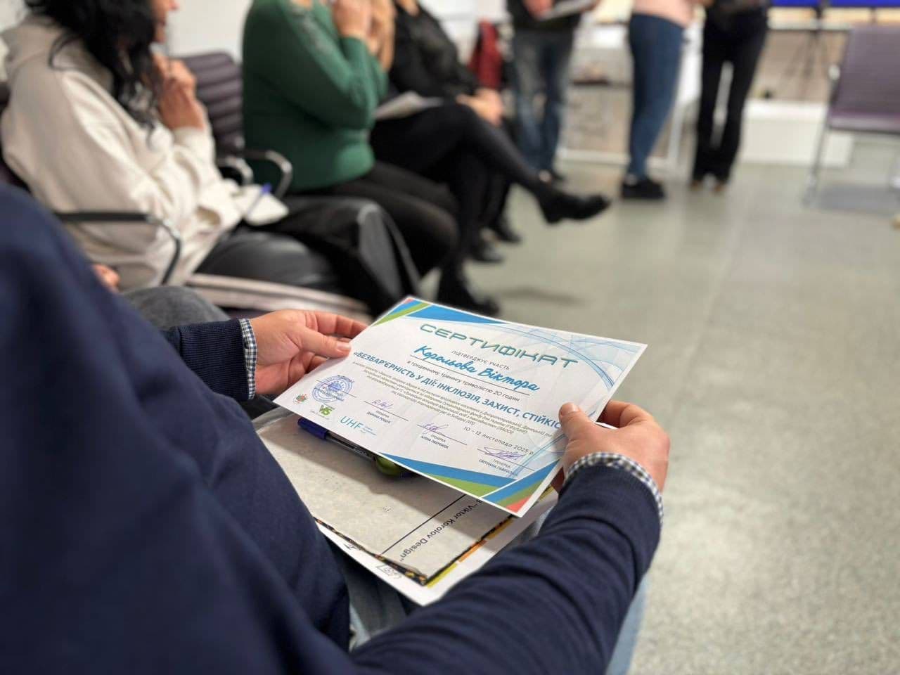 A seminar participant holds a certificate awarded for attending a training on resilience, psychological protection, and emergency response. People are seated in a classroom-like setting. The certificate displays the name Karas Viktor, the date November 15, 2023, and organizational logos.