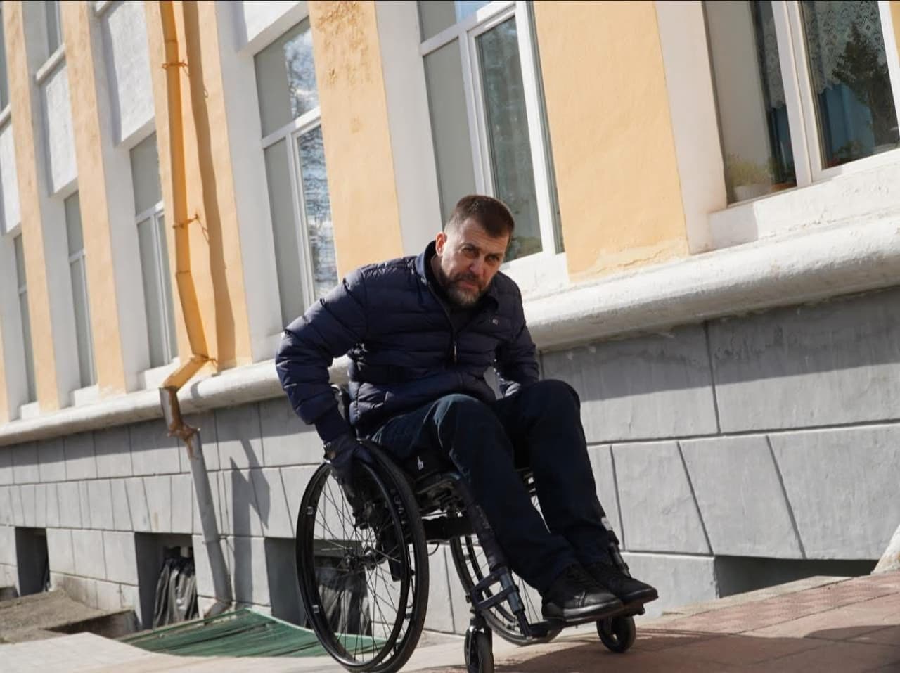 На зображенні людина в інвалідному візку рухається по похилому тротуару біля будівлі з великими вікнами та фасадом жовто-сірого кольору. Вона одягнена в темну куртку та темні штани й докладає зусиль, щоб піднятися вгору. Сцена демонструє виклик для людей на візках у міському середовищі, особливо на нахилених поверхнях.