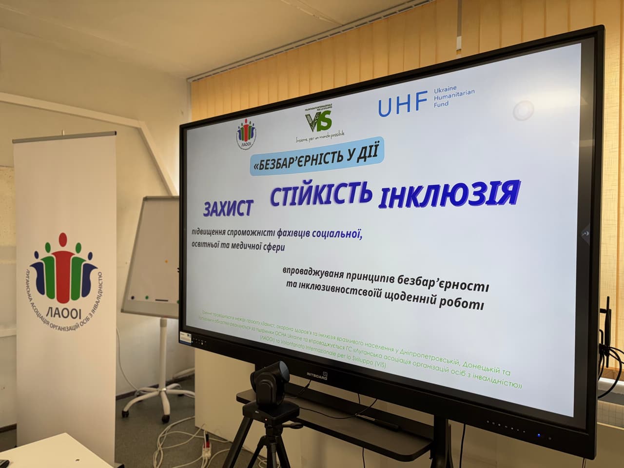 «Екран із презентацією тренінгу “Безбар’єрність у дії: захист, стійкість, інклюзія” у навчальній аудиторії. Праворуч — великий монітор з титульним слайдом, ліворуч — банер ЛАООІ та фліпчарт.