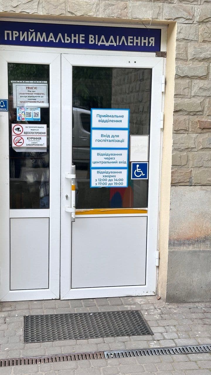 The entrance to a medical facility, specifically the reception department. The glass door displays several informational signs in Ukrainian, indicating hospital services. The signs include directions for hospitalization entry, visiting hours, and accessibility options for disabled individuals. Additionally, there are symbols for video surveillance and a no-smoking policy.