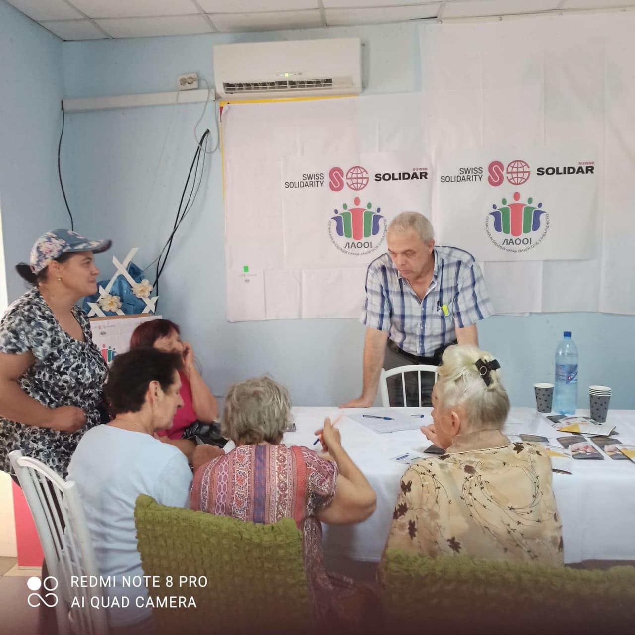 Чотири жінки сидять за столом, одна жінка стоїть, а навпроти біля столу стоїть чоловік.