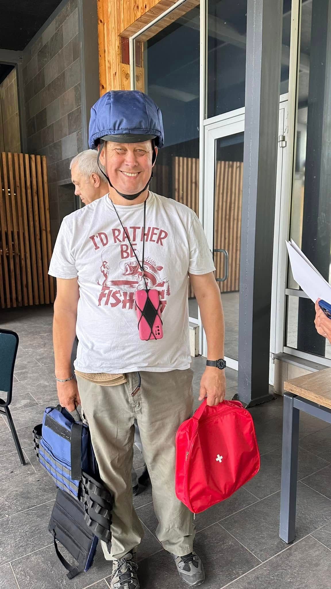 A man is smiling, holding a red, blue, and black bag. Another man is standing behind him.