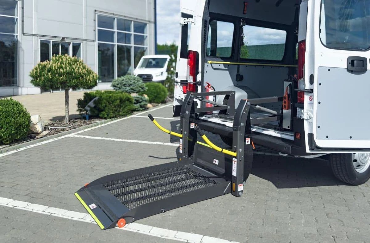A white van with rear doors open is equipped with a wheelchair lift. The platform is extended to the ground, ready for use. Inside, the spacious layout includes handrails designed for accessibility. The van is parked in a lot near a modern building with large glass windows. The scene highlights inclusive transportation solutions for individuals with mobility challenges.
