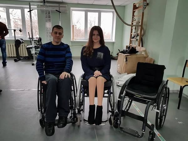 Two young people are sitting in wheelchairs in a room that appears to be a rehabilitation center. The man is wearing a striped sweater and dark pants, while the woman is dressed in a dark dress and black boots. Next to them, there is an empty wheelchair. The room contains exercise equipment, boxes, and wooden shelves. A large window behind them lets in natural light.