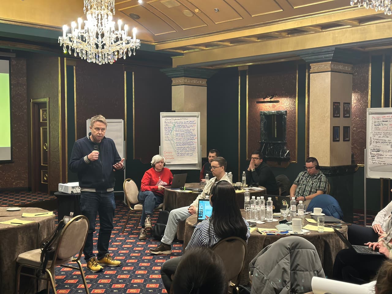 A group of people participating in a roundtable discussion in a conference hall. A man in a wheelchair speaks into a microphone in the foreground.