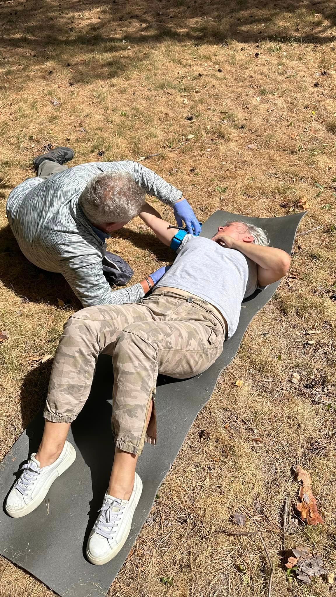 A woman is lying on her back on a mat, while a man nearby, half-lying, is applying a tourniquet to her arm.