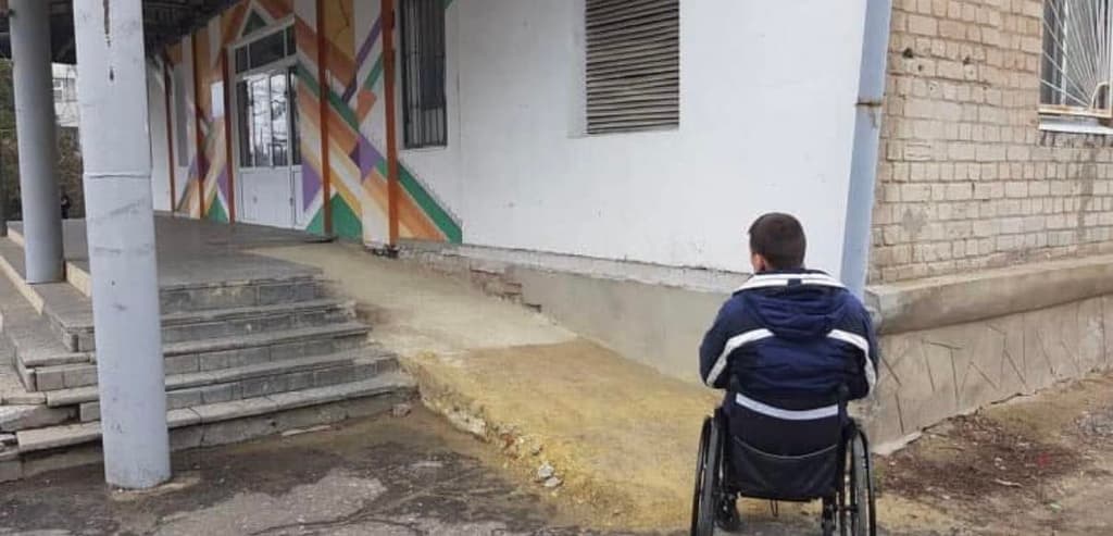 A person in a wheelchair faces a building entrance with colorful geometric wall art. The entrance includes stairs and a steep, uneven ramp that appears unsafe and inaccessible. The image highlights infrastructure challenges for individuals with mobility impairments.