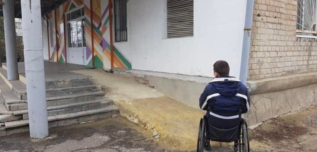 A person in a wheelchair faces a building entrance with colorful geometric wall art. The entrance includes stairs and a steep, uneven ramp that appears unsafe and inaccessible. The image highlights infrastructure challenges for individuals with mobility impairments.