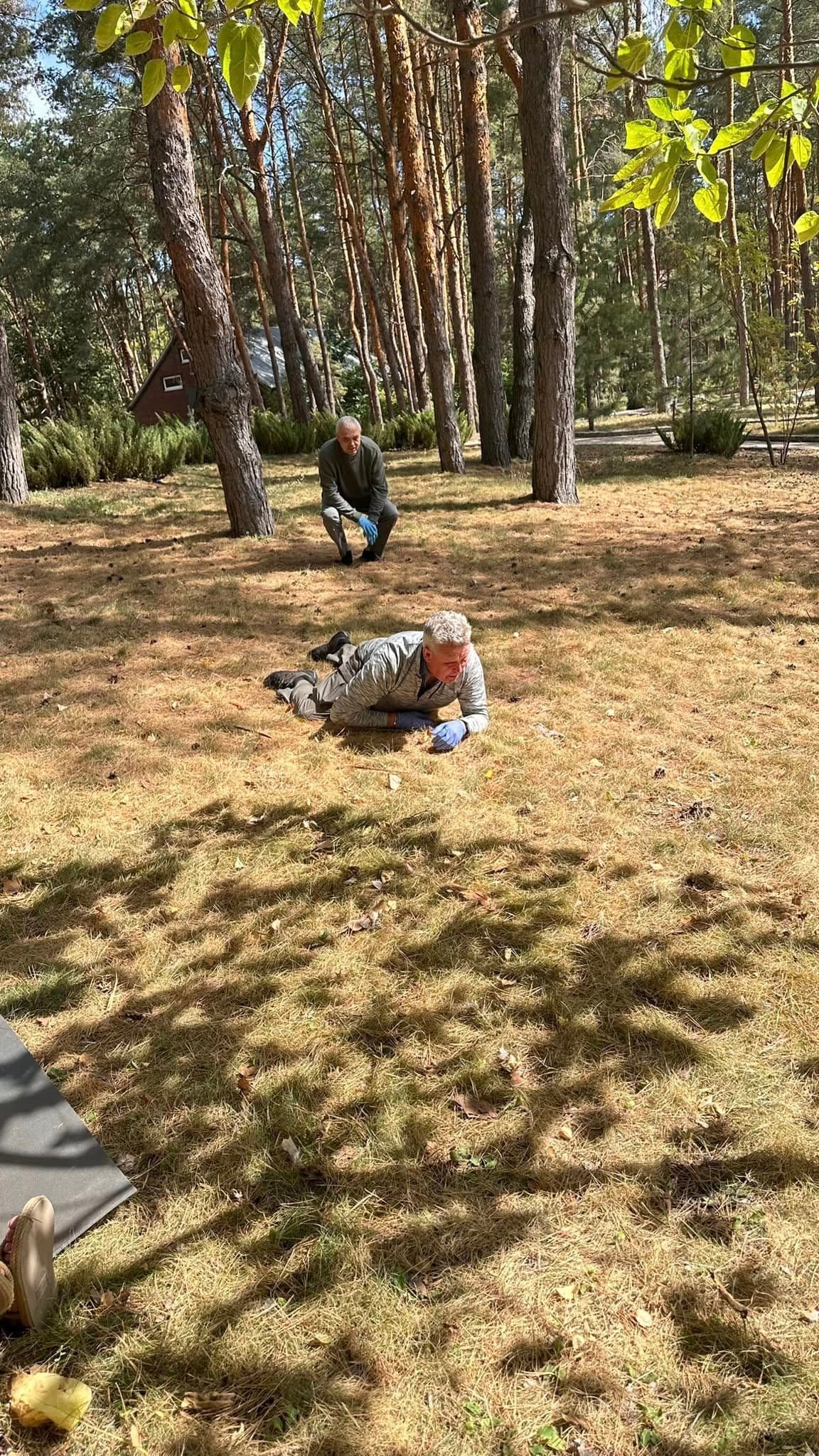 Against the backdrop of a forest and a building, one man is crawling while another crouches, observing.