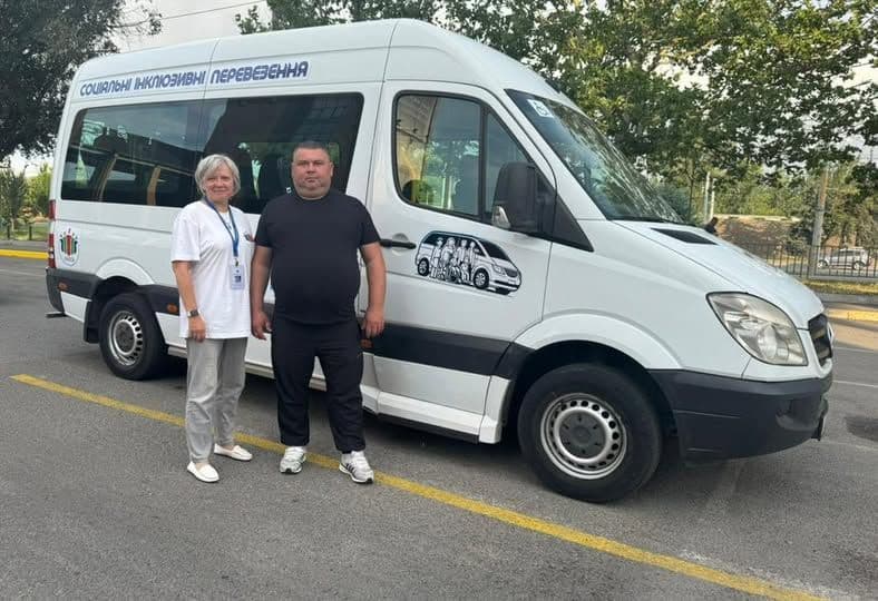 Two individuals stand outdoors next to a white van marked with Cyrillic text reading “СОЦІАЛЬНІ ІНКЛЮЗИВНІ ПЕРЕВЕЗЕННЯ” (Social Inclusive Transportation). An illustrated graphic of a van with passengers appears on the side of the vehicle. The van is parked in a lot, with trees and a metal fence in the background. The individuals are positioned on a yellow painted line marking the space, suggesting an accessible mobility initiative in a residential or community setting.