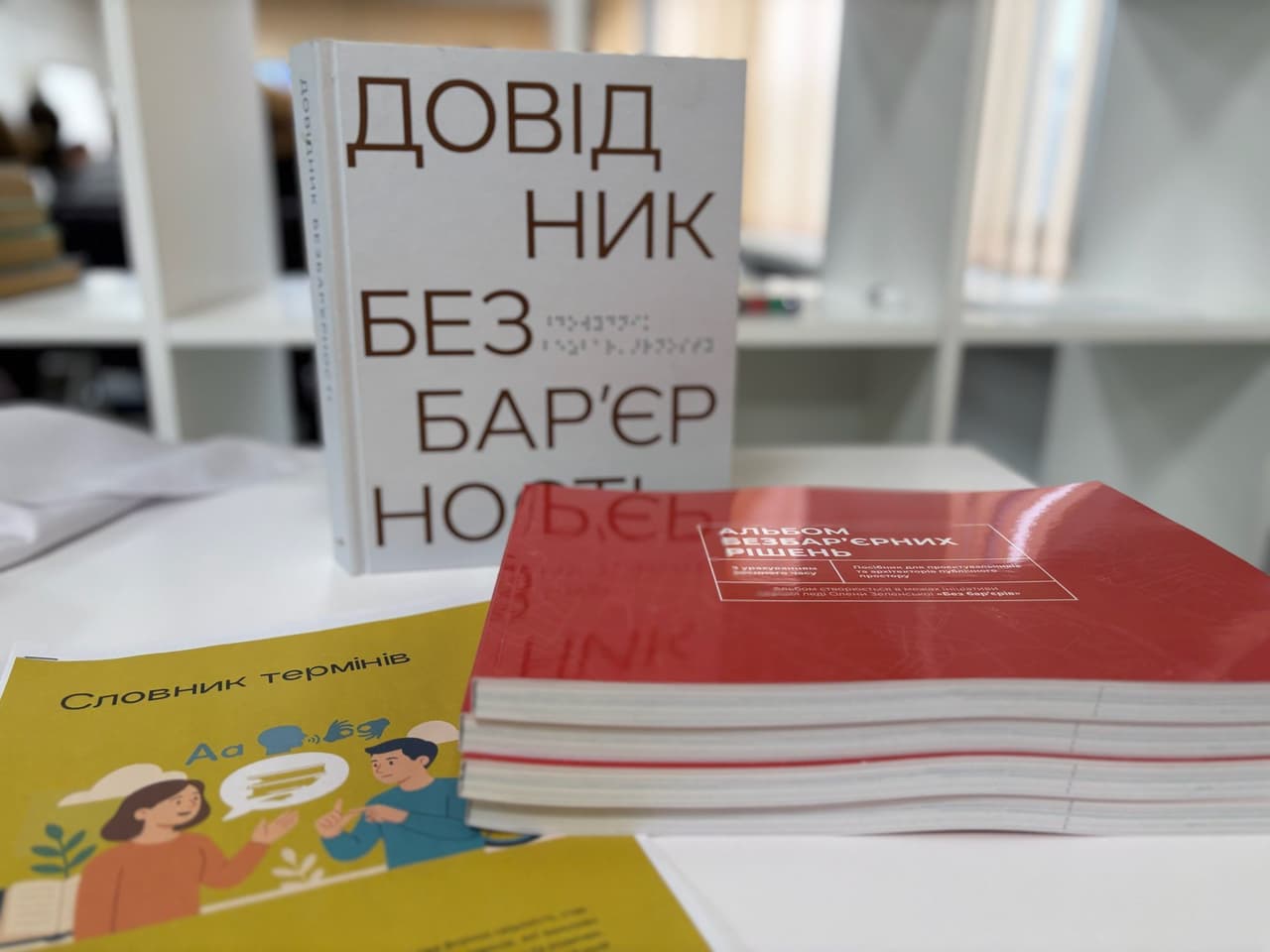 На столі в сучасному приміщенні розкладені українськомовні інформаційні матеріали: біла книга з написом і шрифтом Брайля «Довідник без бар’єрів», червоні буклети «Альбом безбар’єрних рішень» та жовта брошура «Словник термінів» з ілюстрацією двох людей. Матеріали присвячені темі доступності та інклюзивного спілкування.