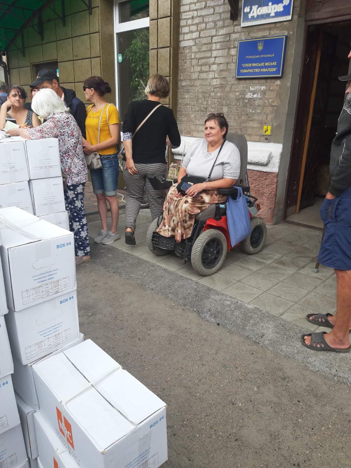 🇬🇧 A woman in an electric wheelchair in front of the building of the public organization “Slovyansk City Society of Disabled People.” Nearby, volunteers distribute humanitarian aid in white boxes with the logo of an international organization. People stand in line, waiting for their turn to receive assistance.
