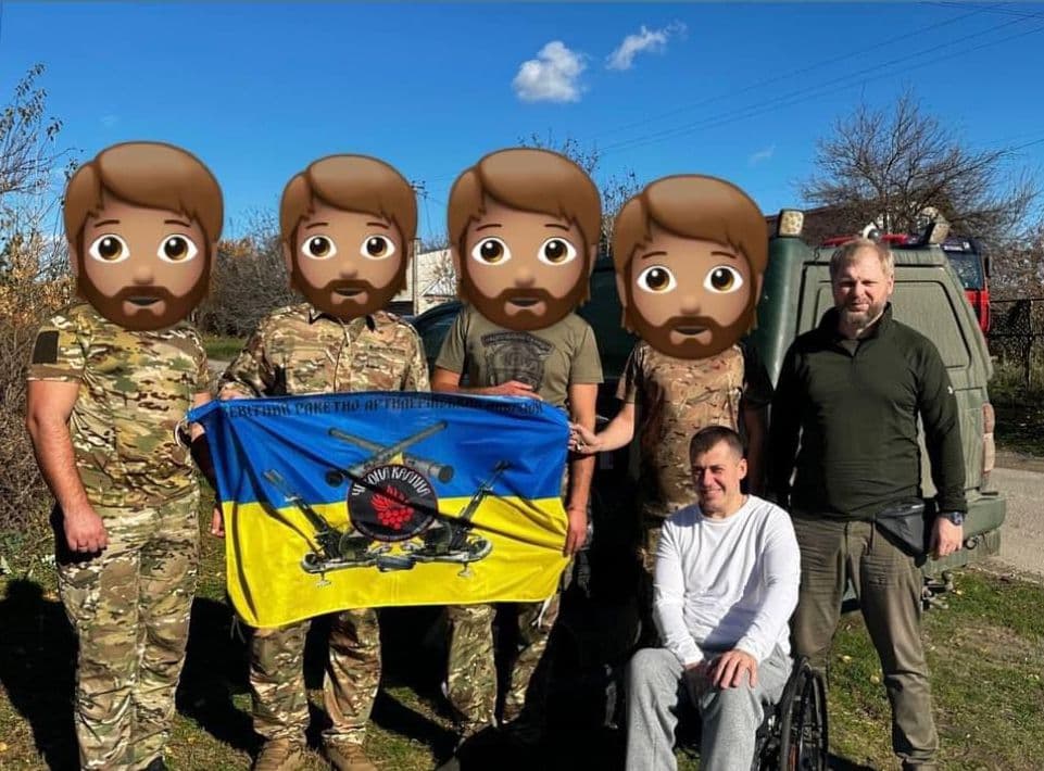 Четверо військових із закритими обличчями тримають прапор, поруч стоїть ще один чоловік, а один чоловік сидить в інвалідному візку.