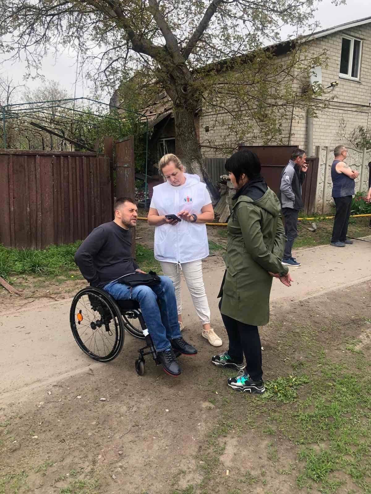 Зображення показує групу людей на свіжому повітрі біля будинку. Одна людина знаходиться у інвалідному візку, інша носить білий халат і тримає телефон, а третя стоїть поруч у зеленій куртці. У фоні видно ще двох людей: одна стоїть зі схрещеними руками, а інша з руками в кишенях. Сцена розташована у сільській або приміській місцевості, на фоні видно ґрунтову дорогу, дерев'яний паркан і зелену рослинність. Здається, що присутні взаємодіють або ведуть розмову.