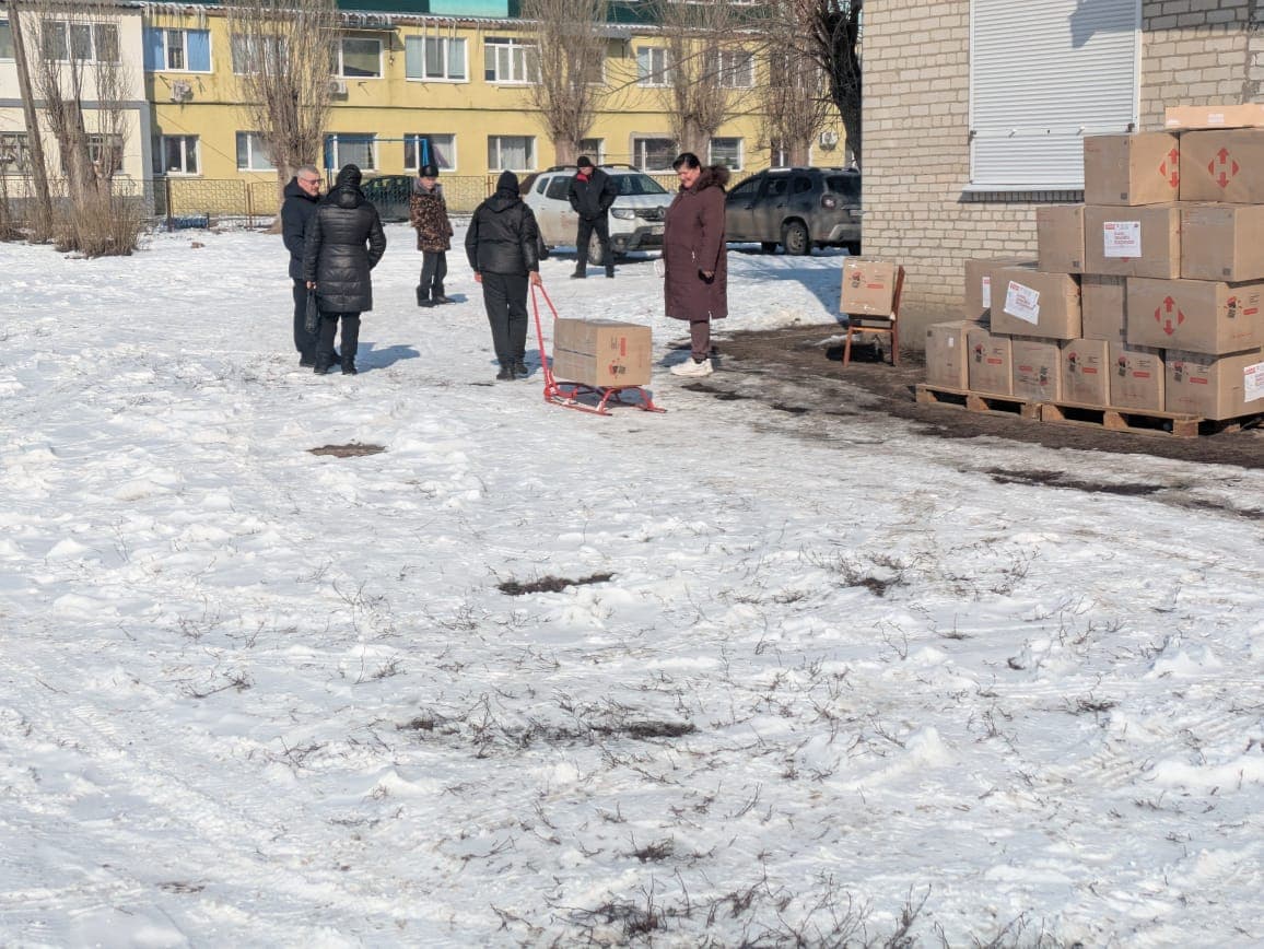 A large number of humanitarian aid boxes are stacked near a building. A car with an open trunk is ready for transport.