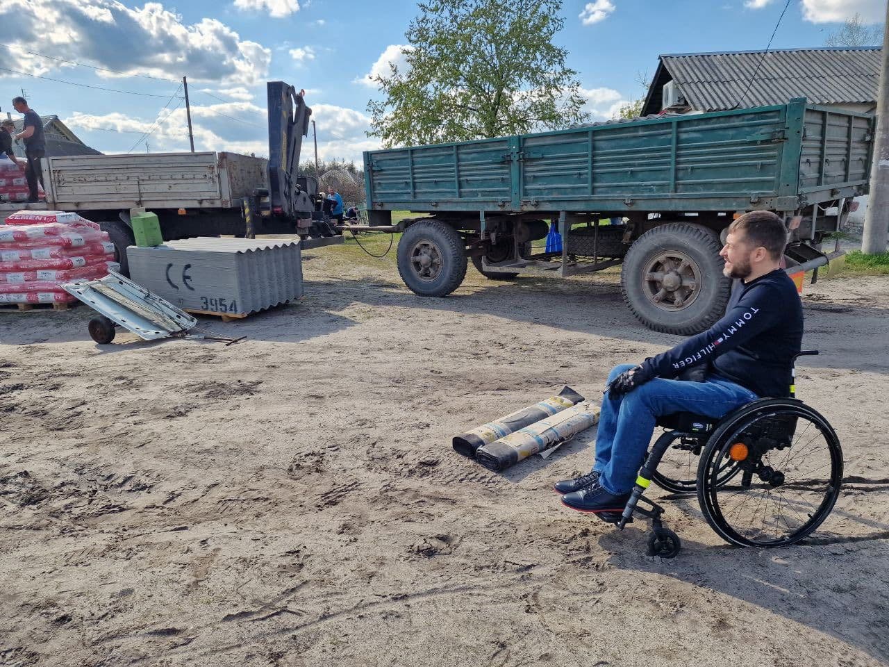 A man in a wheelchair observes an outdoor worksite. He wears a black long-sleeve shirt with “TOMMY HILFIGER” on the sleeve and blue jeans. In the background are two trucks—one loaded with red and white bags, the other green with large tires. Nearby are metal panels, a container marked “CE 3954,” two cylindrical objects, and a small cart. The scene reflects the involvement of a person with mobility impairments in a construction or logistics setting.