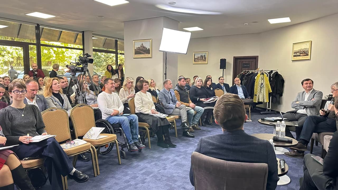 Учасники публічної дискусії сидять у залі, уважно слухаючи виступи. На передньому плані видно частину панелі з мікрофонами та пляшками з водою, а на задньому плані — камери для запису події. Серед присутніх є люди в інвалідному візку. Зал оформлений у нейтральних тонах, на стінах висять картини, а за вікнами проглядається зелень.