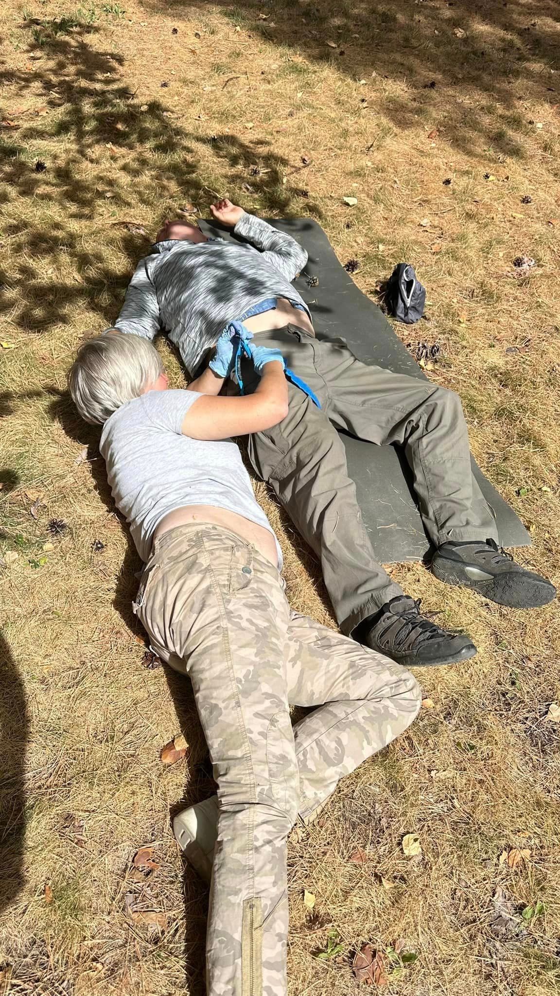 A man is lying on a mat on the grass, while a woman, lying down, is applying a tourniquet to his leg.